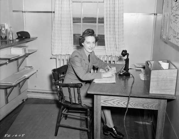 Black and white photograph. A woman sits at a wooden desk. On the desk is paperwork, a small shelf unit, and a microphone. There are wall shelves behind her, and a window to her left. She is in a military uniform.
